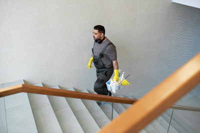 Treppenhausreinigung in Hamburg – Reinigungskraft mit gelben Handschuhen trägt Reinigungsutensilien eine Treppe hinauf.