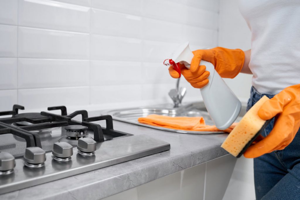 Küchenreinigung in Hamburg – Reinigungskraft mit orangefarbenen Handschuhen reinigt Herd und Arbeitsfläche mit Sprühreiniger und Schwamm.