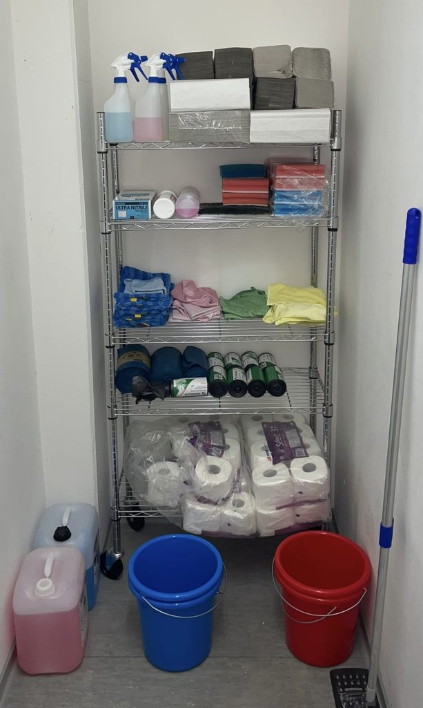 Farblich getrenntes Reinigungsmaterial von Elbreiniger: Tücher und Schwämme im Regal, davor roter und blauer Eimer für Sanitärbereich und Büro